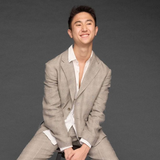 A young man wearing a light gray suit and white shirt sits on a stool against a plain dark background, smiling with his hands clasped between his knees.