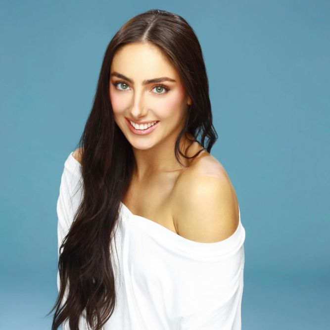 A woman with long dark hair and light eyes smiles at the camera. She is wearing an off-the-shoulder white top and posed against a plain light blue background.