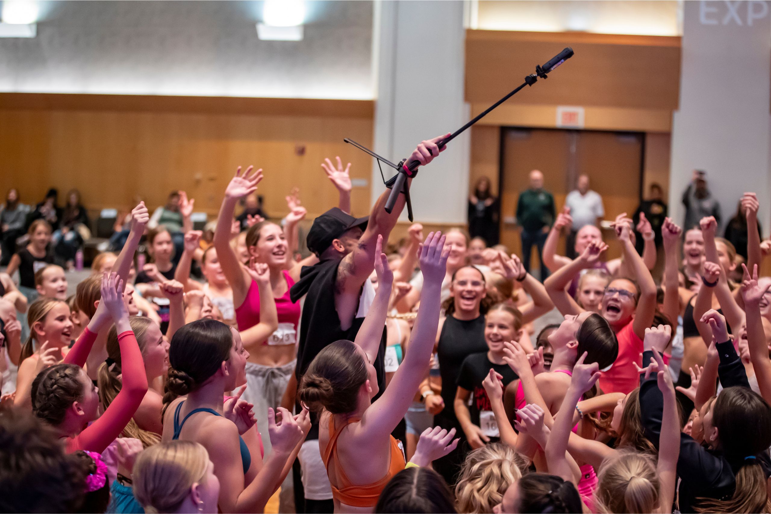 A large group of joyful children and teens in colorful dancewear gather around an adult holding a microphone, cheering and raising their hands in a lively indoor setting.