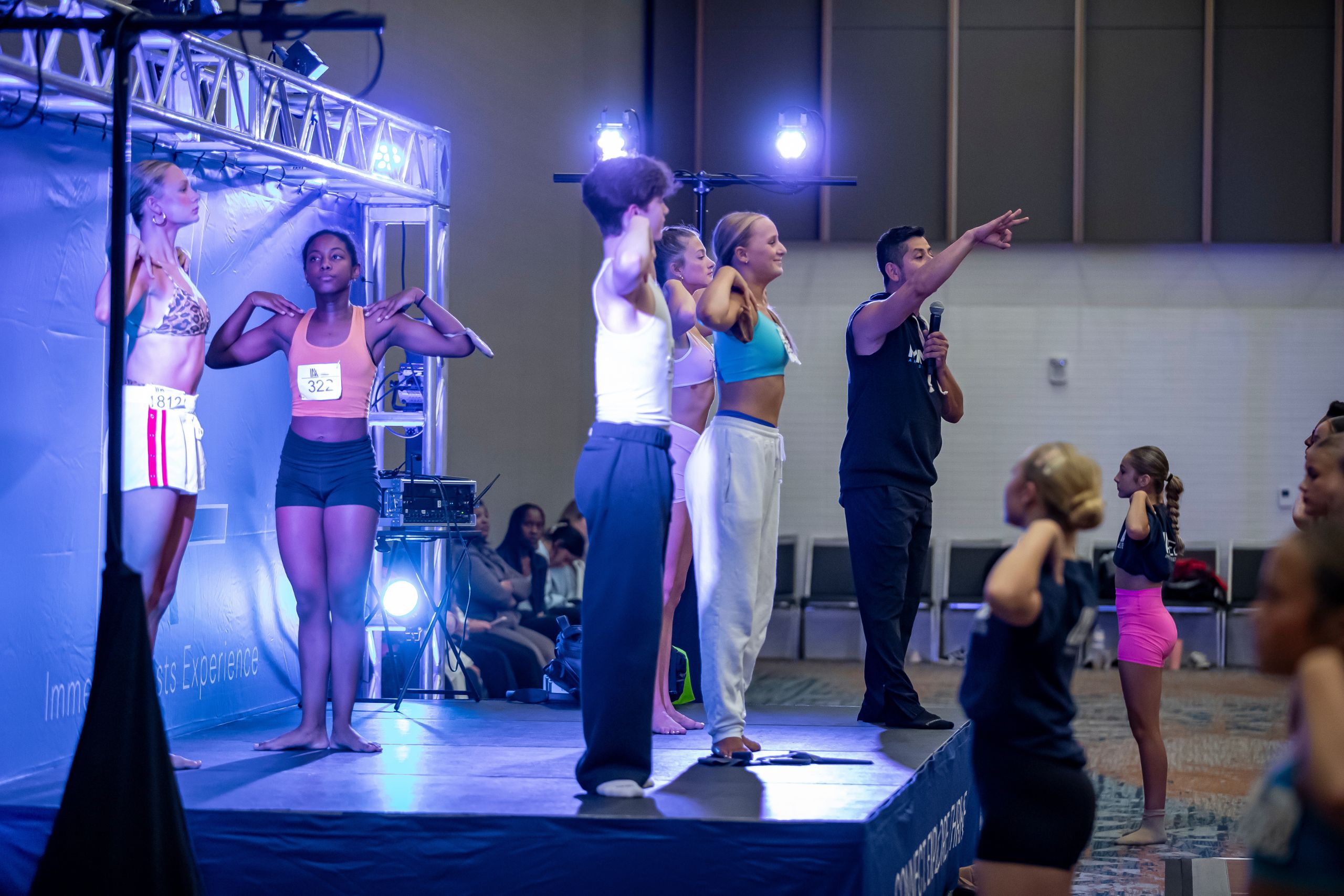 A group of dancers on a stage follow instructions from a person with a microphone. People in workout clothes stand in various poses, with blue lighting and an audience watching in the foreground.