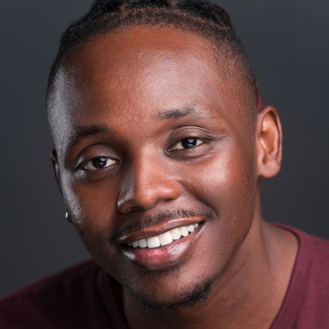 A young man with braided hair smiles warmly at the camera, wearing a maroon shirt against a plain dark background.