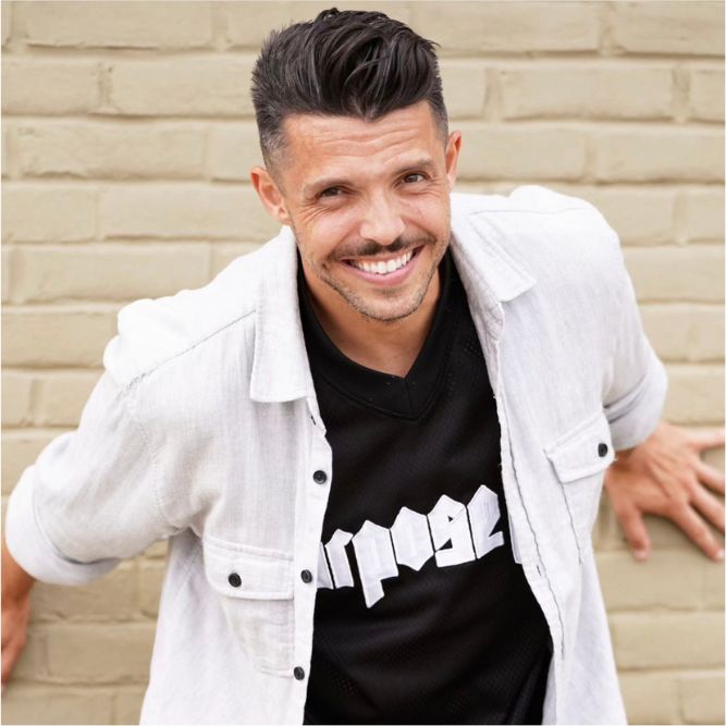 Cedric Botelho - New A man with short dark hair and a trimmed beard smiles at the camera. He is wearing a light-colored button-up shirt over a black t-shirt with white text, standing in front of a beige brick wall.
