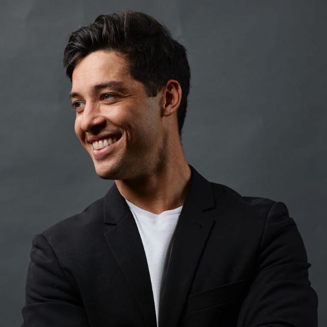 Cole Mills - New A young man with dark hair, wearing a black blazer over a white shirt, smiles and looks to the side against a plain dark gray background.