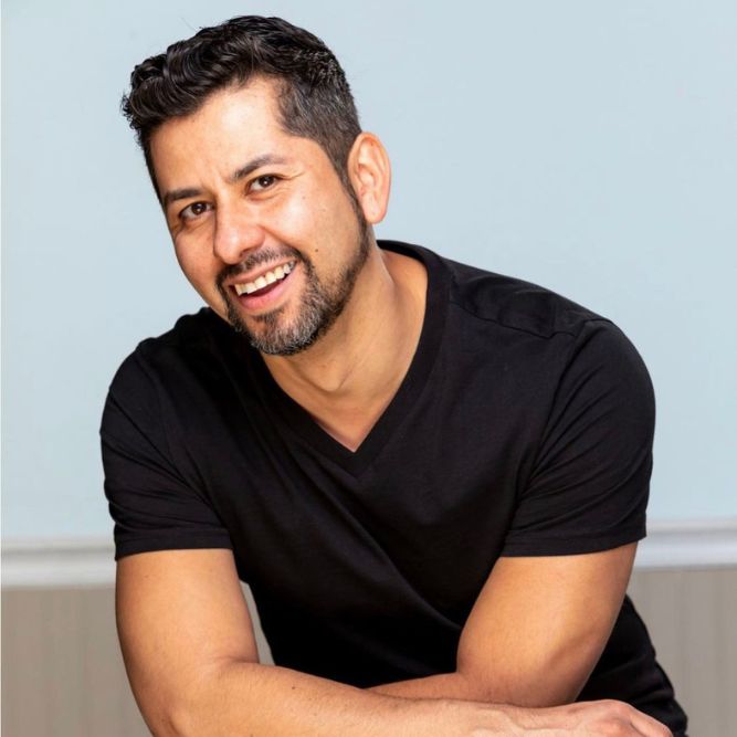 Richard Elszy - New A smiling man with short dark hair and a trimmed beard, wearing a black V-neck t-shirt, sits with his arms crossed in front of him against a plain light blue background.
