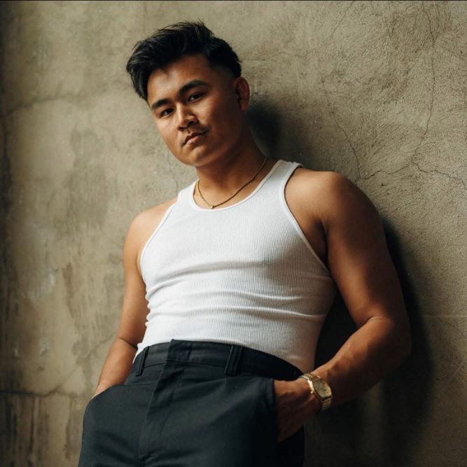 Richy Mon - New A man in a white tank top and dark pants leans against a textured, beige wall, looking at the camera with a relaxed expression. He wears a gold necklace and watch.