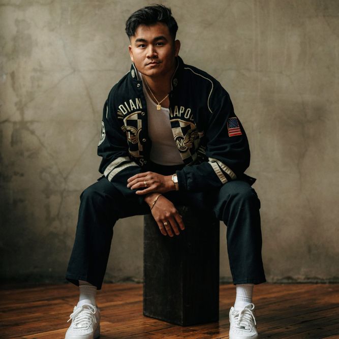 A man with short dark hair sits on a wooden block indoors, wearing a black jacket with patches, black pants, white sneakers, and a white shirt. The background is a textured, beige wall and wooden floor.