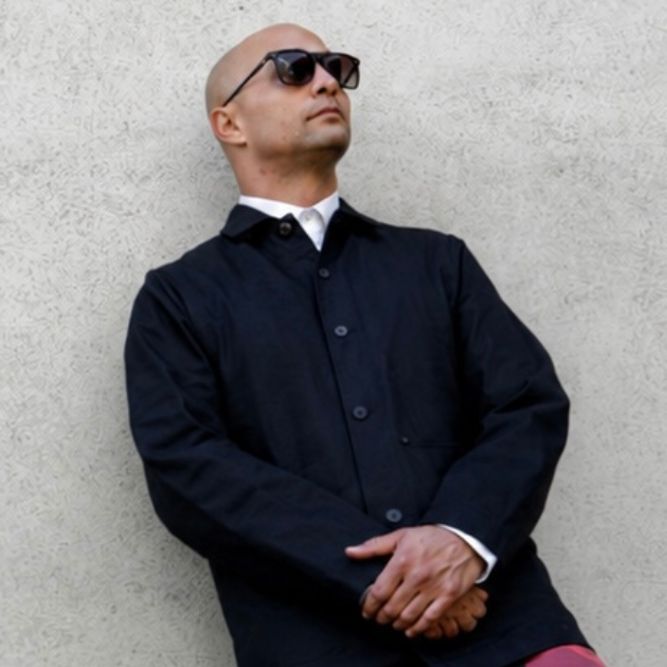 A man with a shaved head, wearing dark sunglasses, a black buttoned coat, and a white shirt, stands against a textured light-colored wall with his hands crossed.