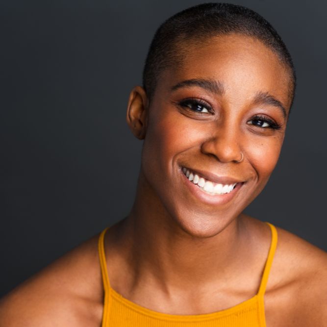 Solace Hough - New A person with short hair smiles warmly at the camera, wearing a mustard yellow sleeveless top, against a dark gray background.