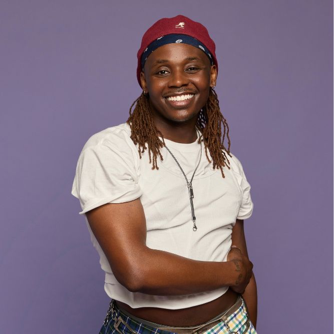 William Harris III - Updated A person with long brown dreadlocks, wearing a maroon hat, white cropped t-shirt, plaid pants, and a necklace, smiles with one arm across their body, standing against a purple background.