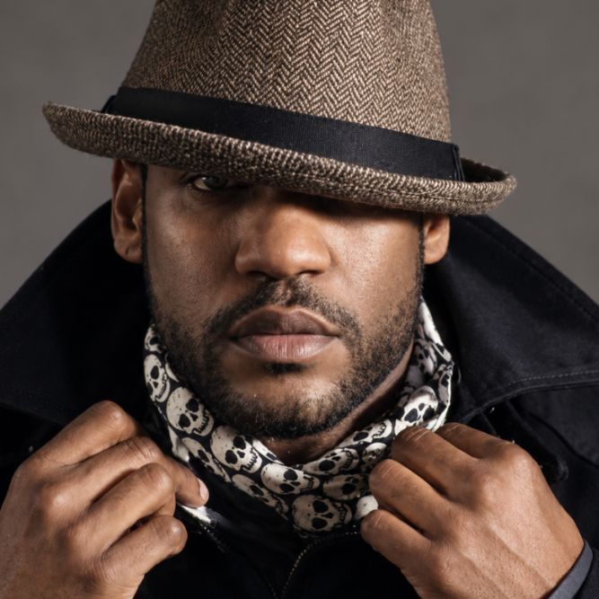 Man wearing a brown fedora, dark coat, and a black-and-white skull-patterned scarf, looking forward with hands gripping his collar. The hat casts a shadow over one eye.